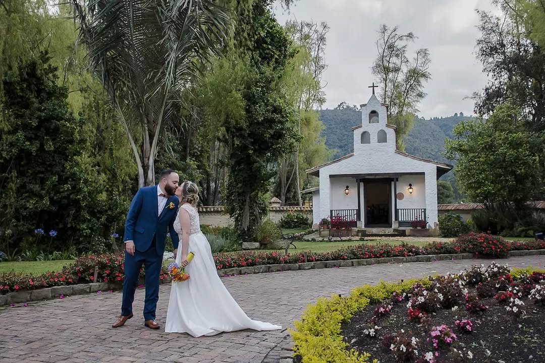 Novios dándose un beso de recién casados en un camino empedrado de la Hacienda Remanso del Río en Cajicá, con la capilla blanca de fondo y jardines florales.